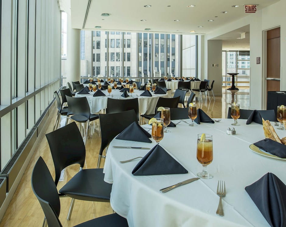 Large, circular tables surrounded by chairs line bight floor-to-ceiling walls in a large dining room at the Gleacher Center