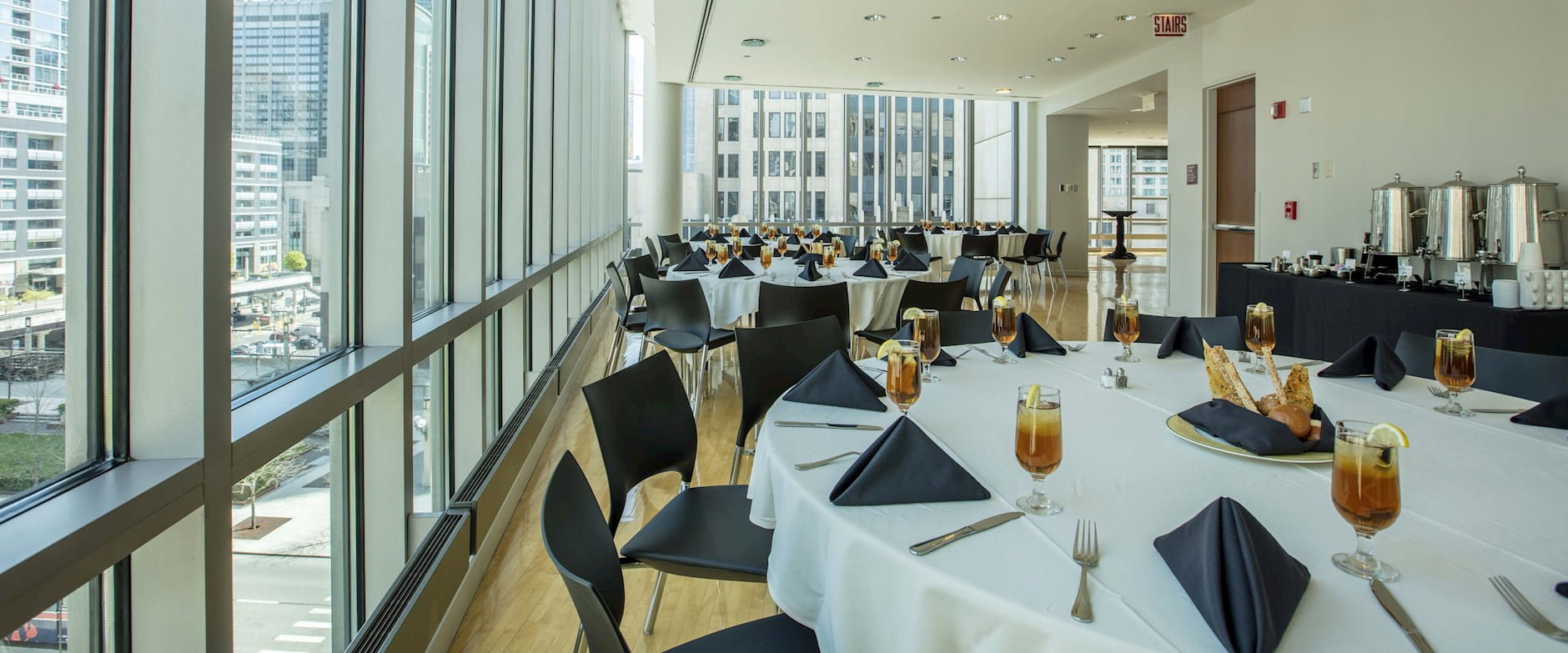 Large, circular tables surrounded by chairs line bight floor-to-ceiling walls in a large dining room at the Gleacher Center