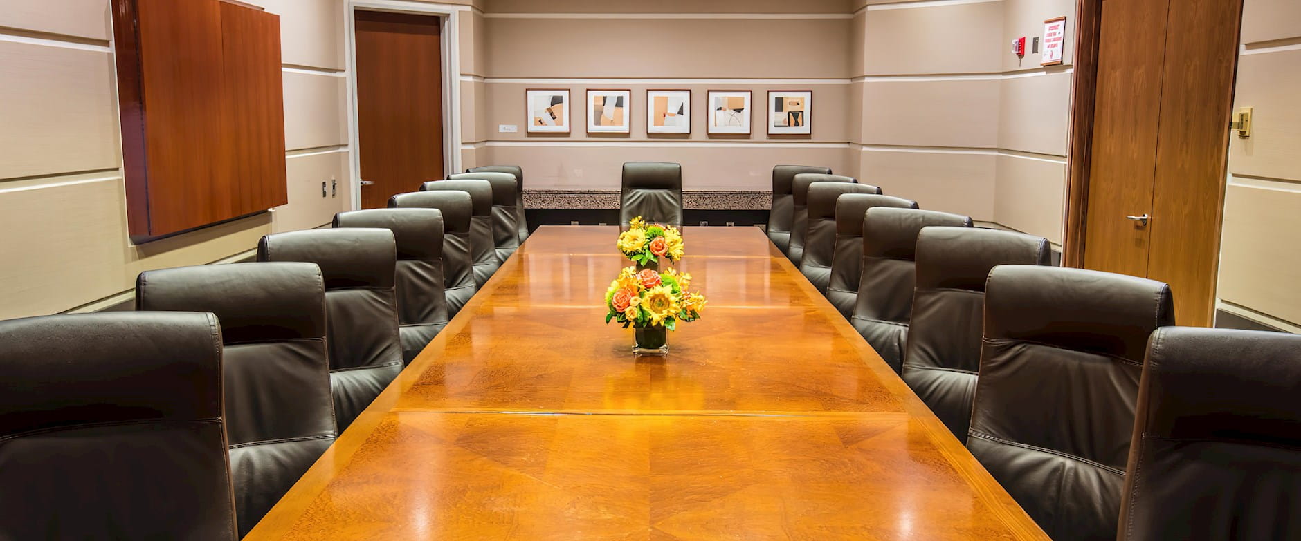 A large, warm wooden boardroom table in the Gleacher Center surrounded by leather office chairs; bouquets of flowers decorate the table