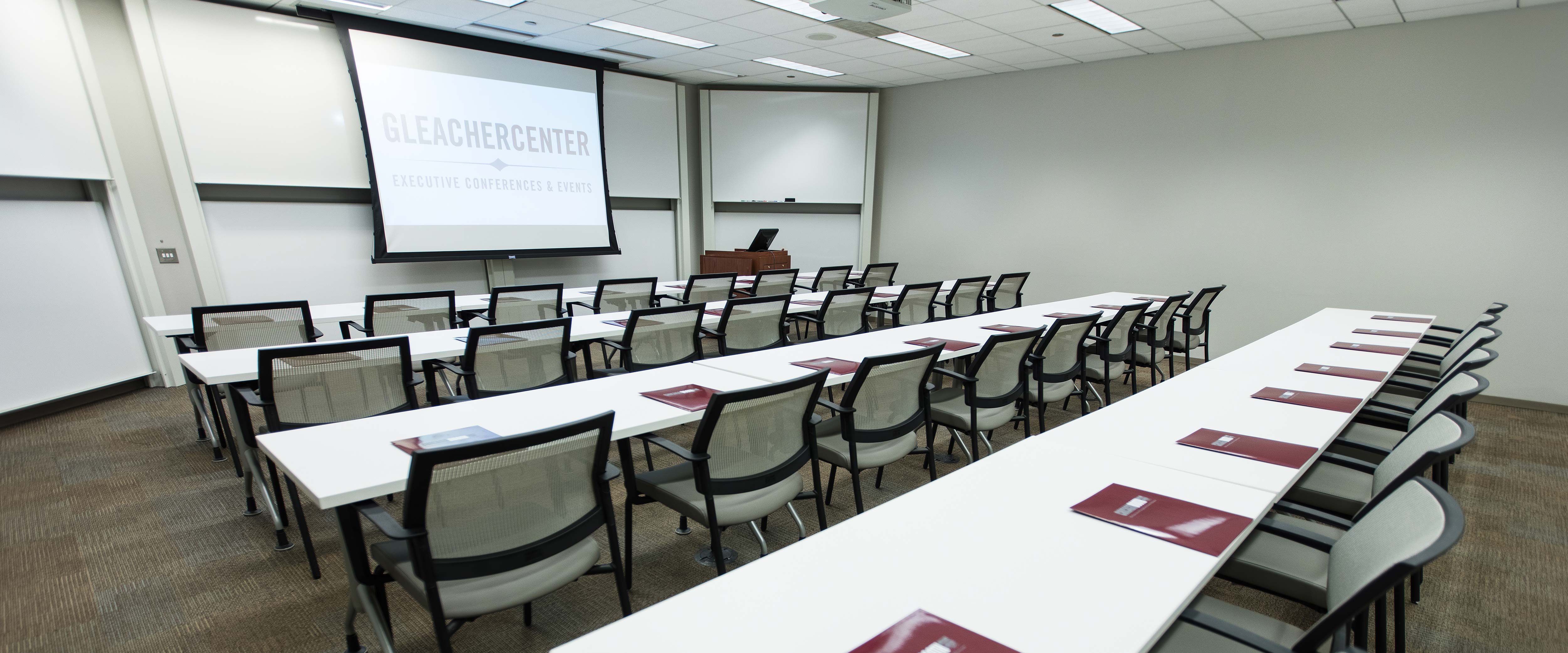 Multi-function room at the University of Chicago Gleacher Center with four rows of tables and chairs facing the projector screen