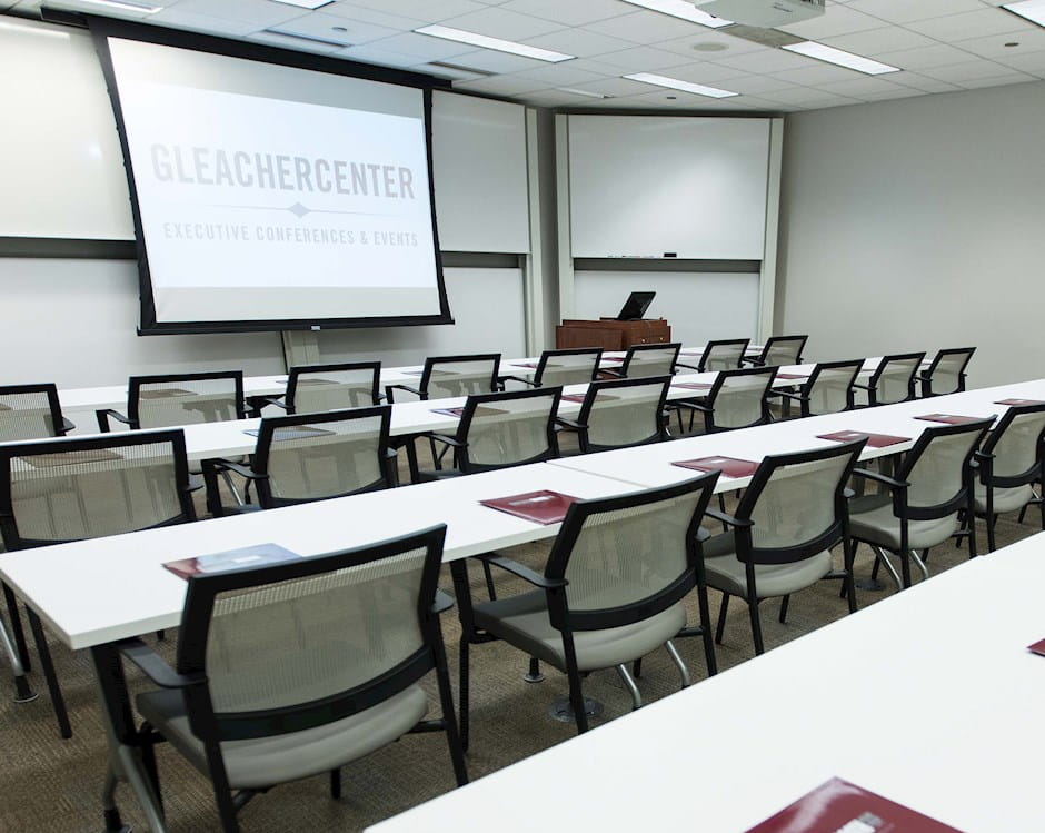 Multi-function room at the University of Chicago Gleacher Center with four rows of tables and chairs facing the projector screen
