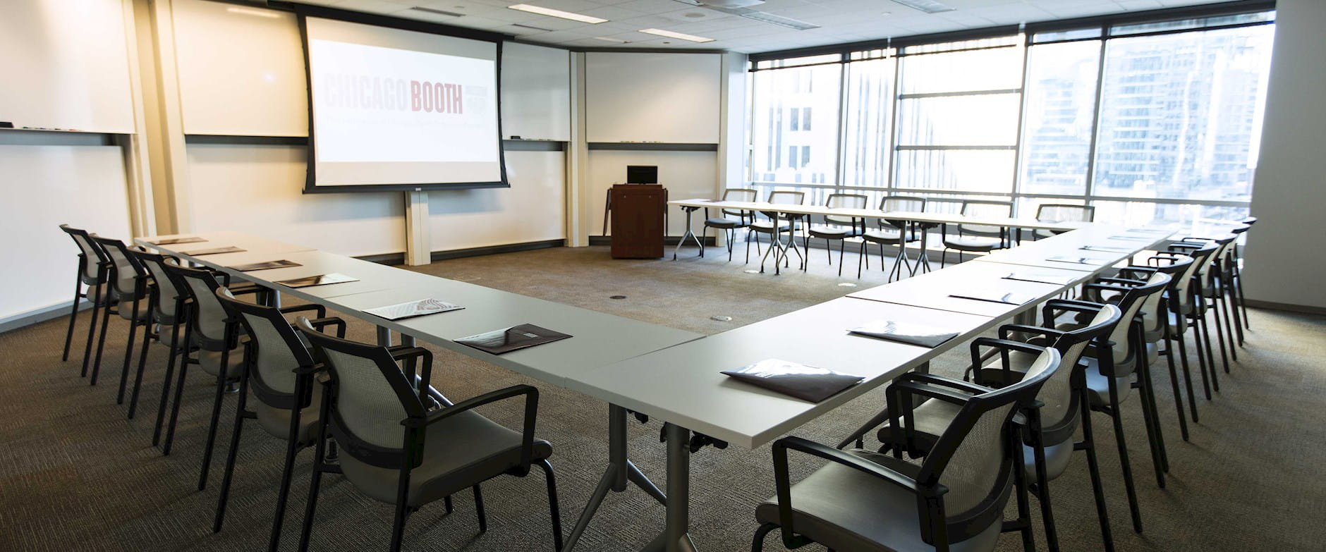 Room 422 in Gleacher Center showing the full semi-circle of desks and chairs with a projector screen