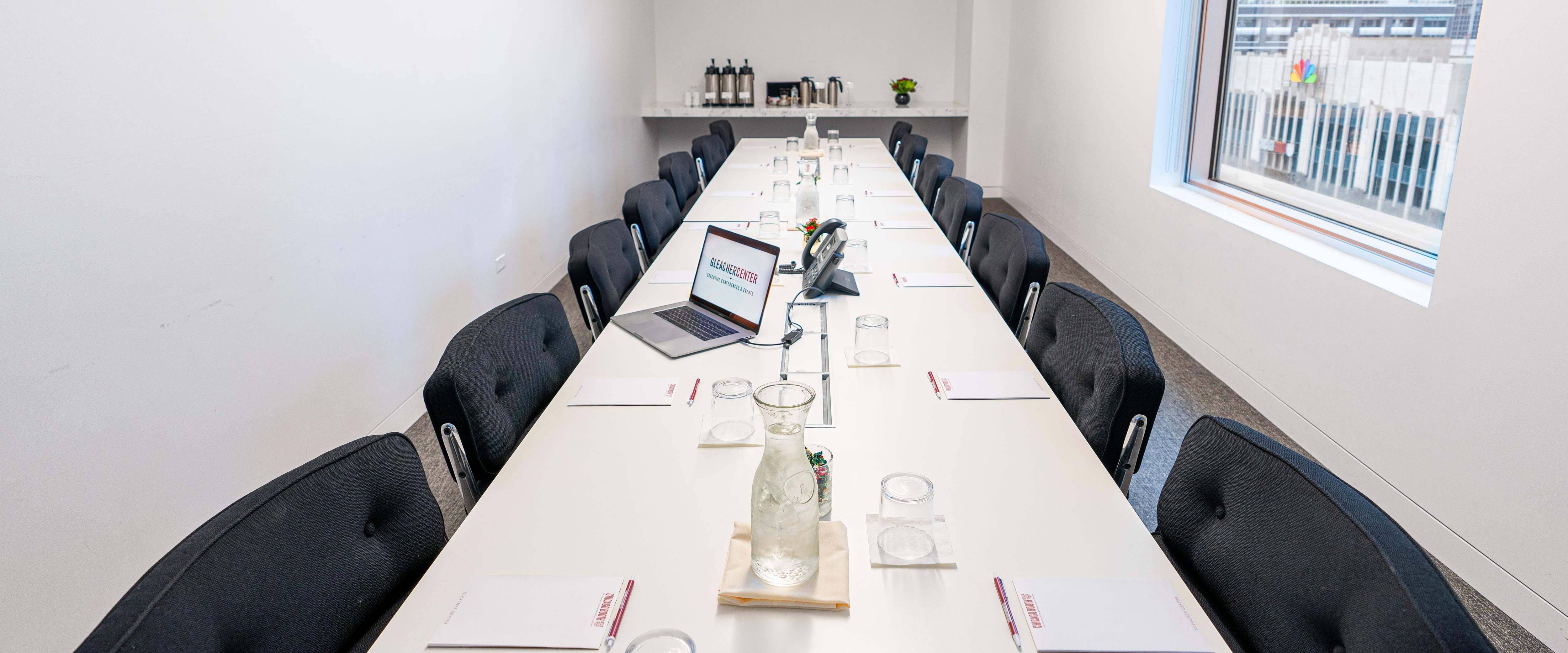 Long boardroom table in the Gleacher Center with drink glasses, notepads, a laptop, and phone