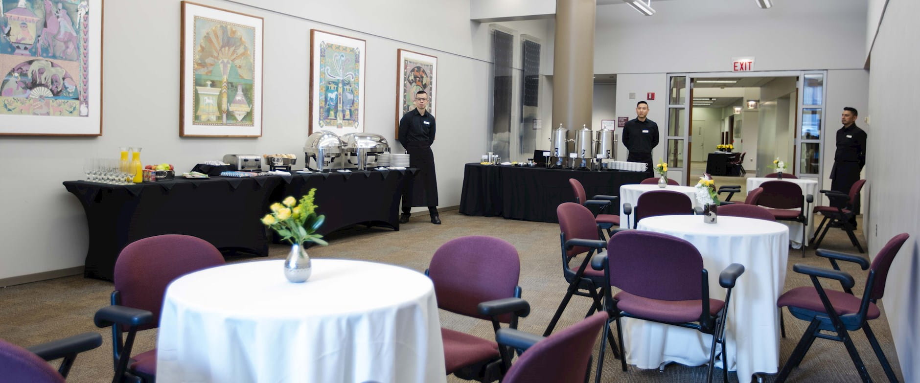 The lobby area of room 100 at the Gleacher Center with a snack buffet and coffee station set up at the back; 3 service staff pose in the back with round tables and chairs in the front