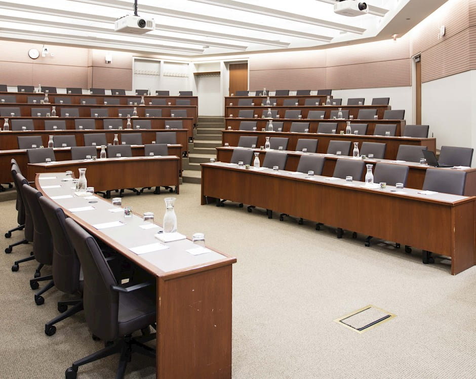 View from the front-left of room 100 at the Gleacher Center showing tiered tables and chairs in a semi-circle with a podium and presenter's seat at the front