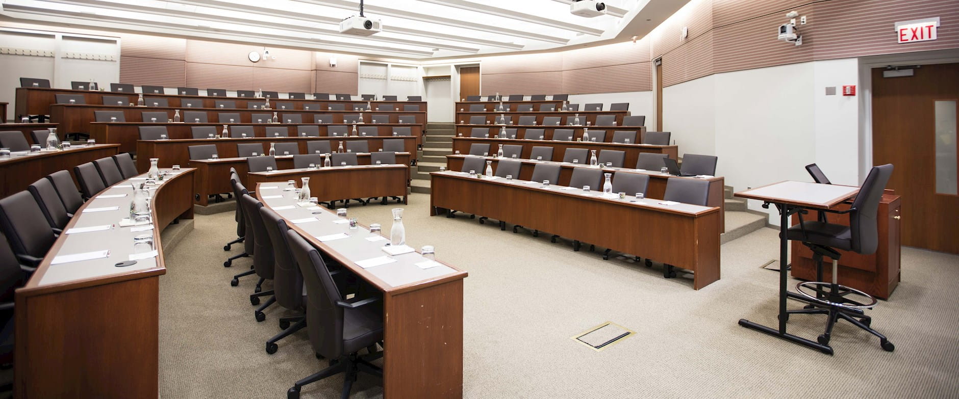 View from the front-left of room 100 at the Gleacher Center showing tiered tables and chairs in a semi-circle with a podium and presenter's seat at the front