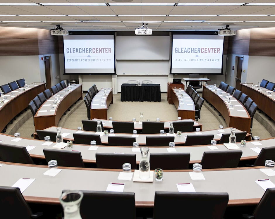 View from the back of room 400 at the Gleacher Center looking out over tiered tables and chairs in a semi-circle with a presenters' table and 2 projector screens at the front