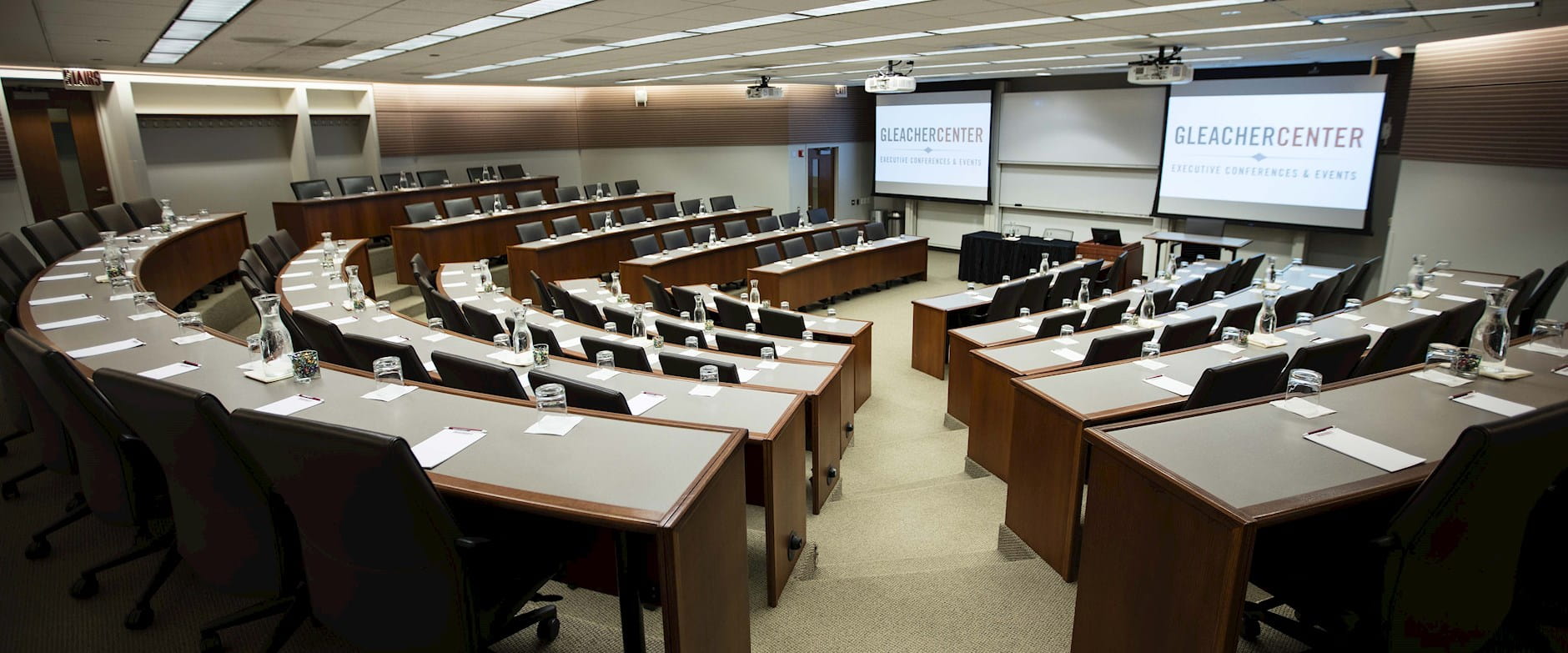 A wide view from the back of room 400 at the Gleacher Center showing tiered tables and chairs in a semi-circle with 2 projector screens at the front